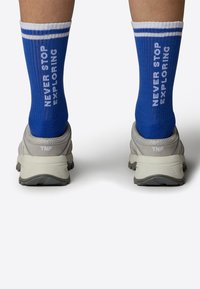Niebieskie skarpetki w białe paski z napisem "NIGDY NIE PRZESTAWAJ EKSPLOROWAĆ". Noszone z szarymi butami trekkingowymi, które mają teksturowane gumowe podeszwy.