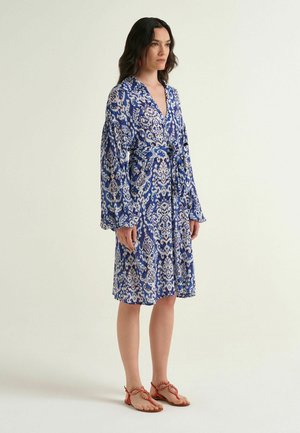 Donna che indossa un vestito a fantasia blu e bianco fino al ginocchio con maniche lunghe e vita annodata, abbinato a sandali arancioni intrecciati.