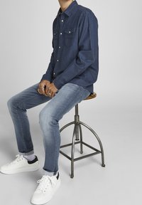 Marindenskjorta i denim med spetsig krage och två bröstfickor, kombinerad med ljusblå jeans och vita sneakers, sittande på en metallpall.