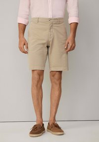 Beige Baumwollshorts mit flachem Bund, Seitentaschen und einem ein Knopfverschluss. Tragen mit braunen Wildlederschuhen und einem hellrosa Hemd.