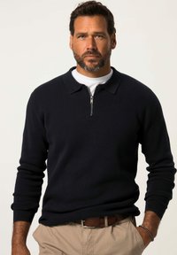 Dunkelblauer Strickpullover mit einem Reißverschlusskragen, strukturiertem Stoff und langen Ärmeln. Kombiniert mit khakifarbenen Hosen und einem braunen Gürtel.