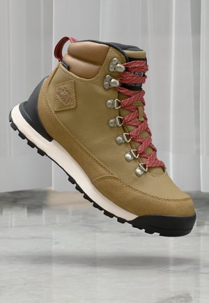 Beige turstøvler med en teksturert overdel, brune lærdetaljer, røde prikkete snører og en svart gummisåle. Har metalløyler og en pull-tab.