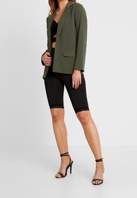 Blazer verde oliva su un crop top nero, abbinato a pantaloni corti neri con bordi in pizzo. Tacchi alti neri con cinturini completano l'outfit.