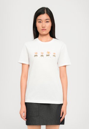 Femme portant un T-shirt blanc avec quatre fleurs roses et jaunes et le texte "PS Paul Smith", associé à une jupe gris foncé.