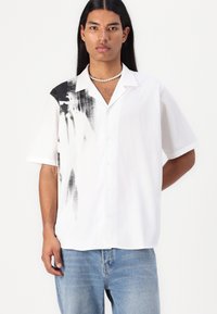Camicia bianca a maniche corte con bottoni, caratterizzata da una stampa astratta nera sulla spalla sinistra. Realizzata in tessuto leggero, vestibilità rilassata.