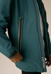 Veste imperméable turquoise avec poches latérales zippées et des détails jaunes contrastants, ainsi que des poignets velcro. Texture lisse avec un design moderne.