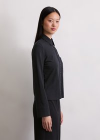 Camicia nera a maniche lunghe con bottoni e colletto, taschino frontale e taglio dritto; realizzata in tessuto morbido con una texture liscia.