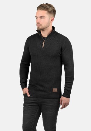 Hombre con cabello corto y barba, vestido con un suéter negro texturizado con cuello de media cremallera y pantalones oscuros, de pie frente a un fondo liso.
