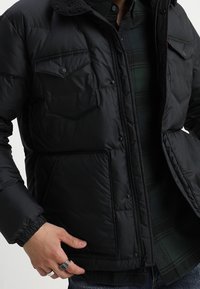 Sort pufjakke med blød krave, trykknap-låg lommer og elastiske manchetter. Båret over en mørkternet skjorte. Glat tekstur, isoleret design.