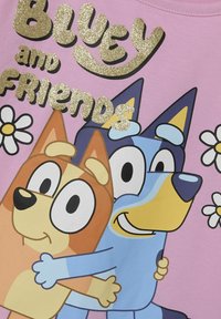 Różowa koszula z dwoma kreskówkowymi psami w pozie przytulania, z wyraźnymi czarnymi konturami i złotym brokatowym napisem "Bluey i Przyjaciele."