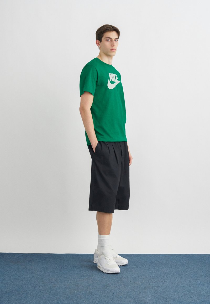 Junger Mann steht mit Händen in den Taschen, trägt ein grünes Nike-T-Shirt, schwarze knielange Shorts, weiße Socken und weiße Turnschuhe auf blauem Teppich.