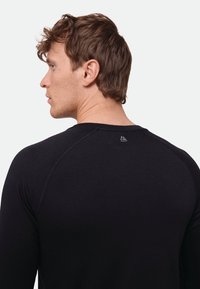 Camiseta deportiva negra de manga larga con tela texturizada, cuello redondo y detalle sutil de logo en la parte trasera cerca del cuello.