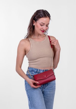 Red synthetic leather crossbody bag with a chain strap, fold-over flap closure, and a subtle logo. Worn with blue high-waisted jeans.