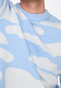 Blauwe en witte gebreide trui met een wolkenpatroon, ronde hals en textuurstof. Close-up van de arm en borstgebied.