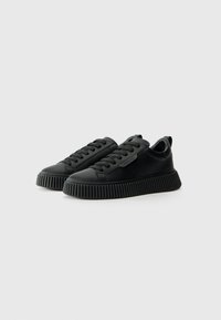 Ein Paar schwarze Low-Top-Sneaker mit dicken, geriffelten Sohlen und schwarzen Schnürsenkeln auf einem schlichten weißen Hintergrund.