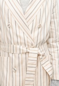 Biely dvojradový blejzer s tenkými hnedými a čiernymi vertikálnymi pruhmi, s opaskom z rovnakého materiálu previazaným v páse cez sivý top.