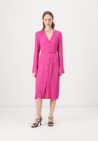 Fuksia midi mekko, jossa on pitkät hihat, V-pääntie, napituskiristys ja vyöllä solmittava nauha. Yhdistetty mustiin korsettikenkiin.