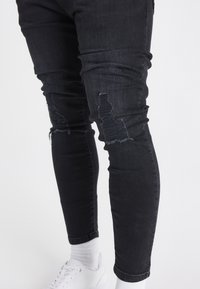Nohy v černých roztrhaných skinny džínách, bílých ponožkách a bílých teniskách na jednobarevném světlém pozadí.