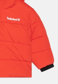 Czerwona pikowana kurtka z przeszytymi sekcjami, z białym logo Timberland. Czarne ściągacze i gładki, lekki materiał.