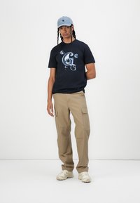 Zwart graphic t-shirt met blauw ontwerp, beige cargobroek en lichtblauwe pet. Witte schoeisel maakt de outfit compleet, staande op een effen achtergrond.