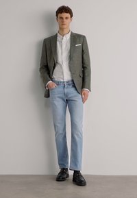 Blazer gri peste o cămașă albă, asortat cu blugi ușor bleumarin și pantofi negri. Materiale sustenabile, croială personalizată, design clasic.