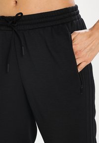 Ruka odpočívající v zipové kapse černých tepláků na stažení s ribbeným pasem a bočním pruhem.