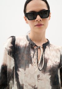 Blusa transparente en tonos claros con patrones florales y rayados. Presenta un escote con lazo y mangas cortas con volantes. Se lleva con gafas de sol negras oversized.