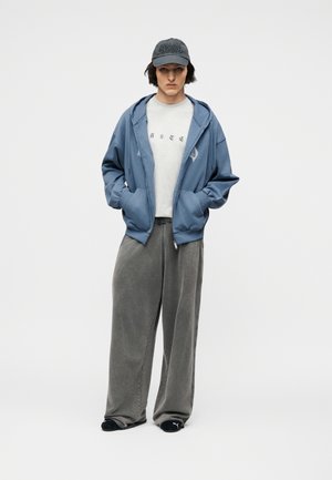 Persoon die een blauwe hoodie met rits draagt over een wit grafisch T-shirt, grijze wijde broek, zwarte Puma slippers en een donkere pet, staand met handen in de zakken.