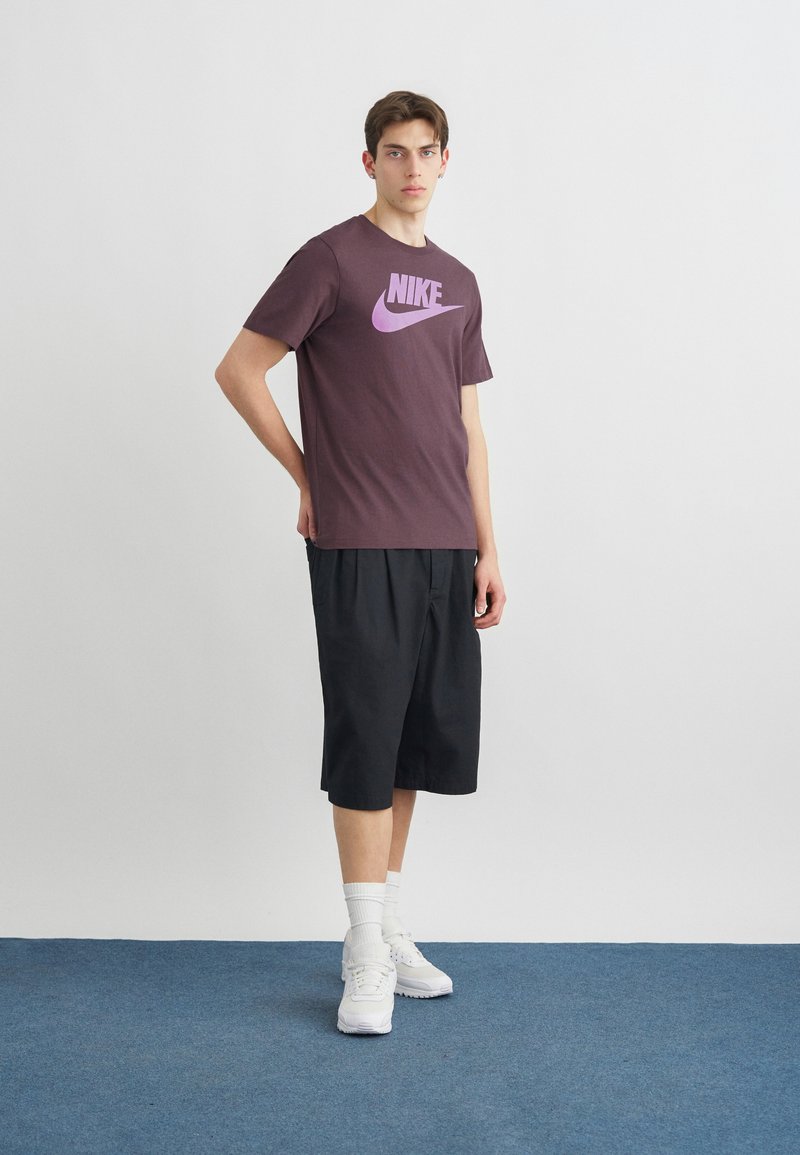 Jonge man staand, gekleed in een paars Nike T-shirt, zwarte wijde shorts, witte sokken en witte sneakers op een blauw tapijt met een effen witte muur als achtergrond.