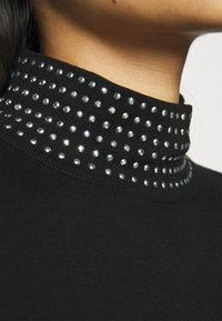 Svart tröja med en hög, ribbad krage som har en rad klara nitar längs kanten. Tyget verkar slätt och åtsittande.