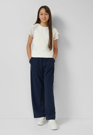 Haut blanc à manches courtes avec des volants superposés, associé à un pantalon large bleu marine et des baskets blanches. Le mannequin pose les mains dans les poches devant un fond neutre.