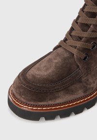 Bota de camurça castanha com superfície texturizada, biqueira redonda e costura clara contrastante. Apresenta atacadores castanhos e uma sola robusta de borracha preta.