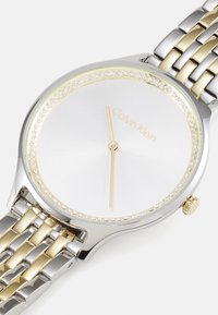 Reloj Calvin Klein con una pulsera de metal en plata y oro, caja redonda, esfera blanca, marcadores de hora dorados y acentos de cristal en el bisel.
