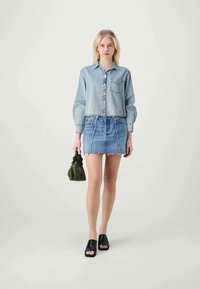Mulher loira a usar uma camisa de ganga clara, uma saia mini de ganga azul, mules pretas de dedo aberto e a segurar uma pequena mala verde-azeitona.