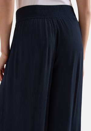 Close-up van een persoon die losse, gestructureerde zwarte broek met elastische tailleband draagt en een wit shirtje in de broek heeft gestopt.