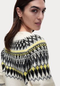 Stickad tröja i krämfärg med svarta och gula geometriska mönster. Har rund halsringning och en mjuk, texturerad yta.