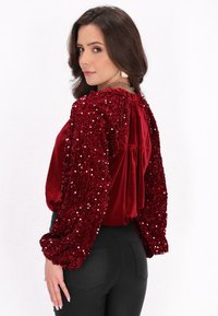 Bluză din catifea roșie cu mâneci bufante, decorate cu paiete, având un design cu spate pliat și o croială lejeră, asociată cu pantaloni negri.