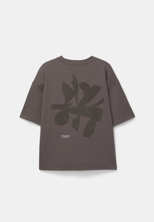 Šedé oversized tričko s krátkymi rukávmi, s veľkým tmavým potlačou na chrbte a malým textovým detailom pri leme.