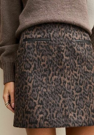 Vrouw draagt een bruine pluizige trui en een rok met luipaardprint met zakken afgezet met zwart kunstleer, hand rust langs haar zij met gouden ringen.