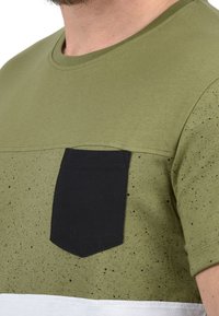 Olijfgroene T-shirt met een zwarte patchzak, spikkelige textuur, ronde halslijn en korte mouwen; witte horizontale streep aan de onderkant.