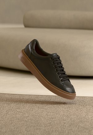 Sneaker in pelle nera con suola in gomma marrone sembra fluttuare sopra un tappeto beige, con un morbido divano beige sullo sfondo.