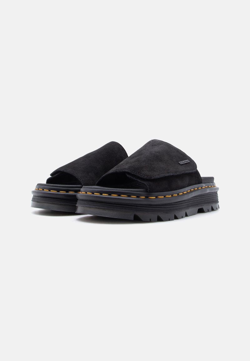 Sandali slip-on in suede nero con suole spesse e robuste e cuciture gialle lungo il bordo della suola su sfondo bianco.