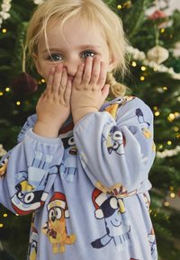 Un maiou de pijama de culoare albastră deschisă, din fleece, cu personaje de desene animate ce poartă căciuli de iarnă. Mâneci lungi cu manșete elastice. Textură moale, design jucăuș.