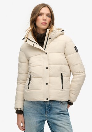 Beige gewatteerde jas met capuchon, ritszakken, zwarte accenten en een gestikt ontwerp. Draagt over een donkere coltrui en blauwe jeans.