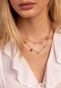 Gelaagde gouden kettingen met bloemen- en bedelpendanten, uitgerust met een gestructureerde ketting en gekleurde kralenaccenten op een lichte achtergrond.
