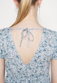 Abito floreale di colore blu chiaro, con una profonda scollatura a V sul retro con chiusura a fiocco, maniche corte gatherate e tessuto testurizzato con piccoli motivi floreali.