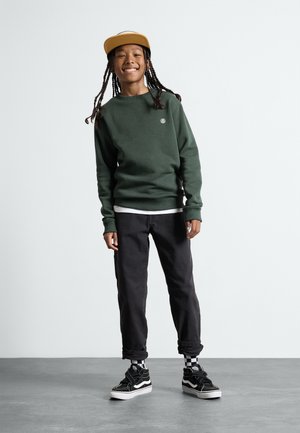 Green sweatshirt, black pants, and black sneakers with white accents. The sweatshirt features a small logo; pants are cuffed at the ankle.