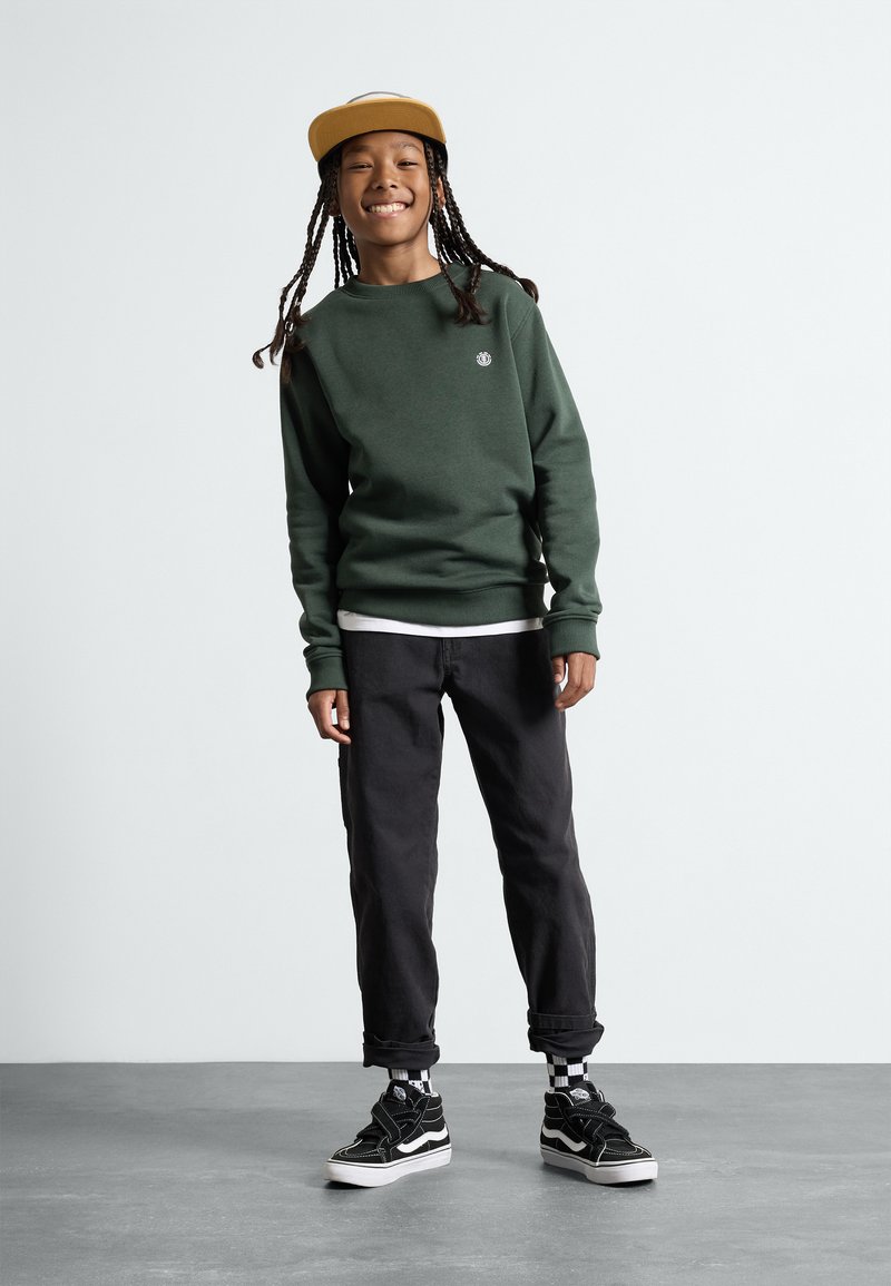 Grüner Pullover, schwarze Hose und schwarze Sneaker mit weißen Akzenten. Der Pullover hat ein kleines Logo; die Hose ist am Knöchel umgeschlagen.