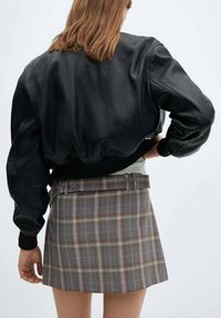Veste bomber en cuir noir avec des manches volumineuses, associée à une mini-jupe à carreaux gris présentant des accents rouges et jaunes, avec un détail de ceinture.