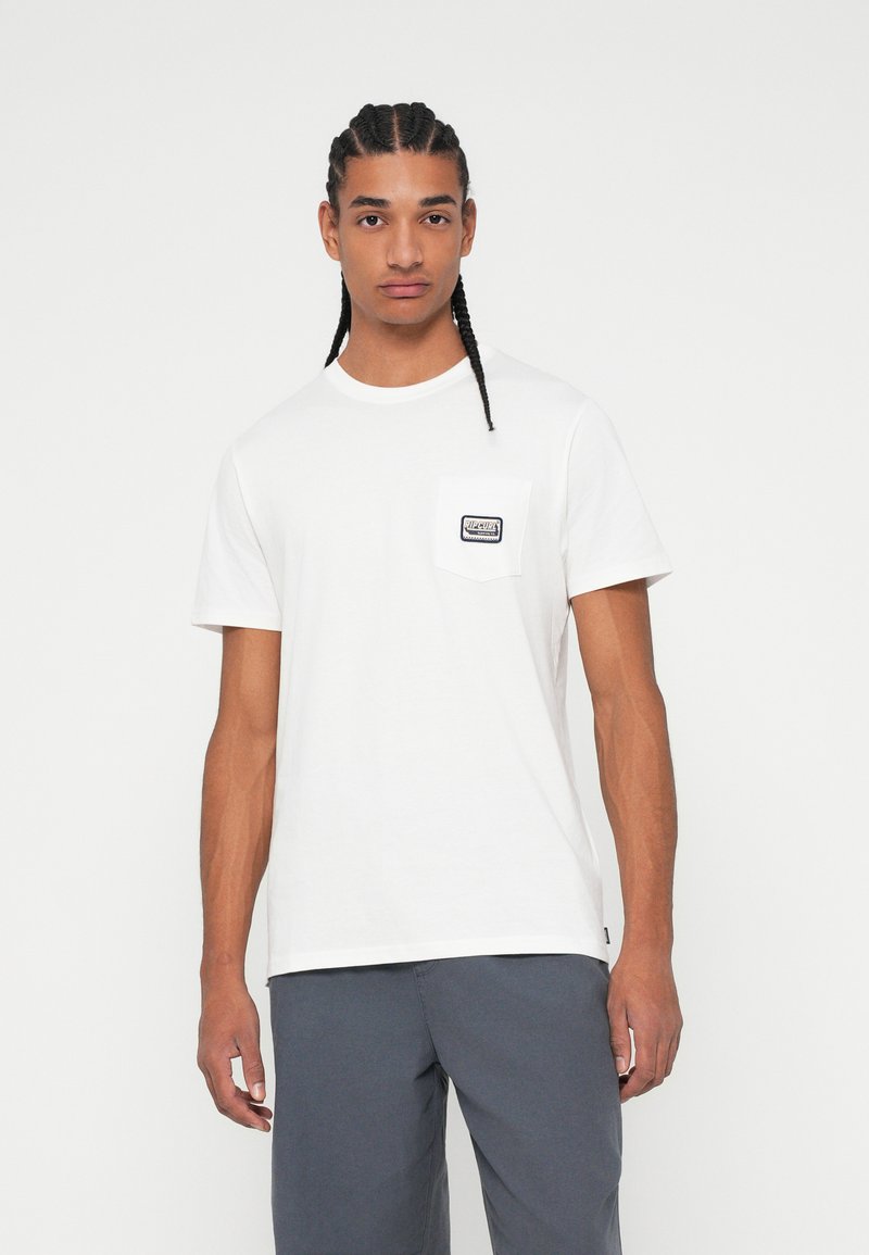 Tricou alb din bumbac cu mâneci scurte, guler rotund și un buzunar pe piept, decorat cu o etichetă cusută. Modelul poartă pantaloni gri.